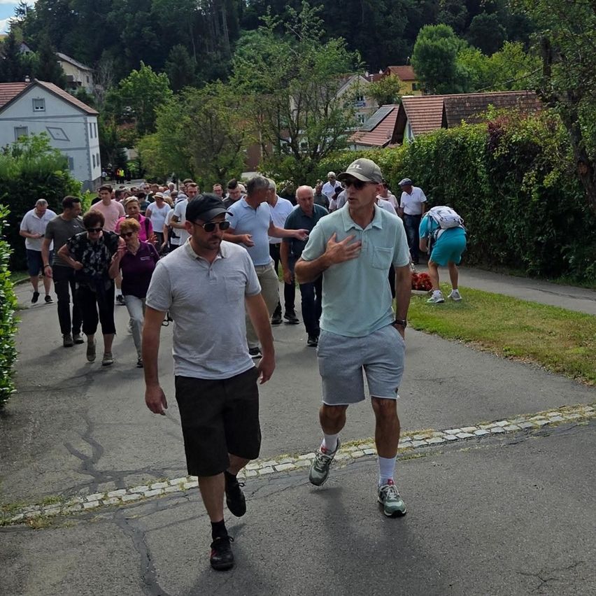 Eine Gruppe von Menschen geht auf einer Straße entlang, die von Häusern und Grünflächen gesäumt ist. Einige tragen Hüte und Sonnenbrillen. Die Straße hat einen Ziegelsteinrand.