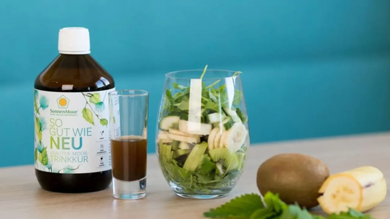 Eine Glasflasche mit Etikett, ein Glas mit brauner Flüssigkeit, eine Glasvase mit grünem Salat und Obst und eine Kiwi auf einem Tisch.
