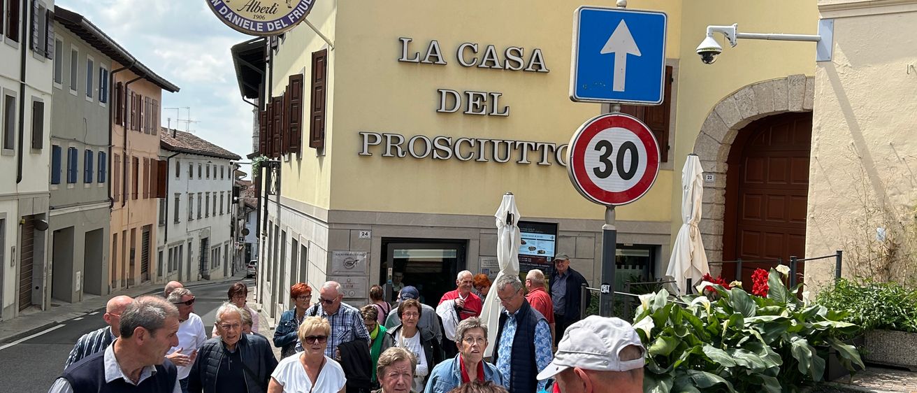 Touristen gehen auf einer Straße an einem Gebäude vorbei, das La Casa Del Prosciutto heißt. Ein blaues Schild mit Pfeil zeigt nach oben, und ein Geschwindigkeitsschild zeigt 30.