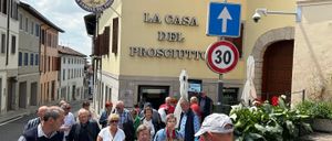Touristen gehen auf einer Straße an einem Gebäude vorbei, das La Casa Del Prosciutto heißt. Ein blaues Schild mit Pfeil zeigt nach oben, und ein Geschwindigkeitsschild zeigt 30.