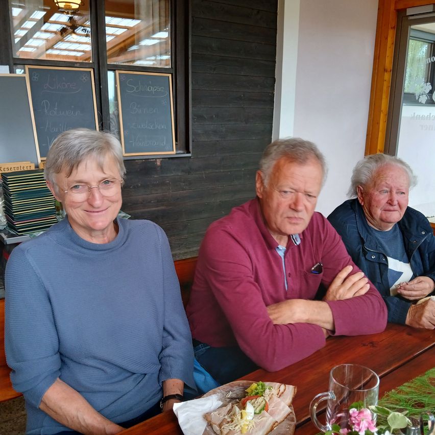 Drei Personen sitzen an einem Holztisch mit Essen. Eine Frau trägt eine Brille und ein blaues Hemd. Ein Mann in einem roten Hemd und ein weiterer älterer Mann in einer blauen Jacke sitzen neben ihr. Hinter ihnen zeigt eine Tafel 'Schlafs' und andere Wörter an.
