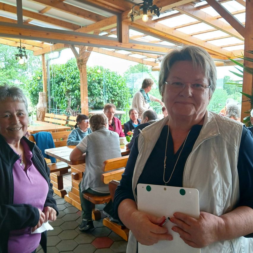 Zwei Frauen stehen in einem Gartenrestaurant, eine hält eine Clipboard mit Stift. Hinter ihnen sitzt eine Gruppe von Menschen an Tischen. Holzstrukturen und Grün umgeben den Bereich.