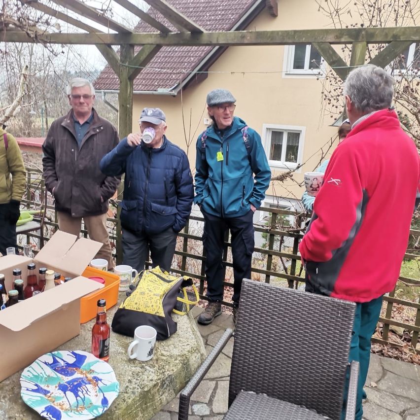 Fünf Männer versammeln sich vor einem Haus, einer trinkt aus einer Tasse. Ein Tisch hält einen Teller, Tassen, Flaschen und eine Tasche. Ein Stuhl steht in der Nähe.