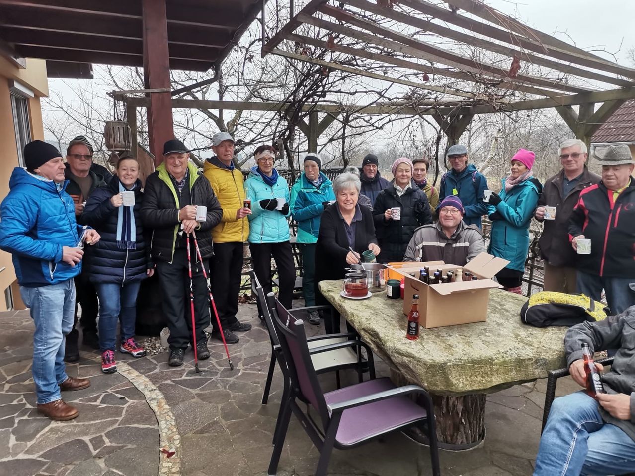 Eine Gruppe von Menschen steht in einem Außenbereich, vielleicht für ein Foto. Sie halten Tassen und einige tragen Mützen und Brillen. Ein Tisch steht vor ihnen mit einer Kiste, Flaschen und anderen Gegenständen. Eine Frau sitzt auf einem Stuhl vor dem Tisch und ein Mann neben ihr. Ein hölzernes Bauwerk steht hinter ihnen.