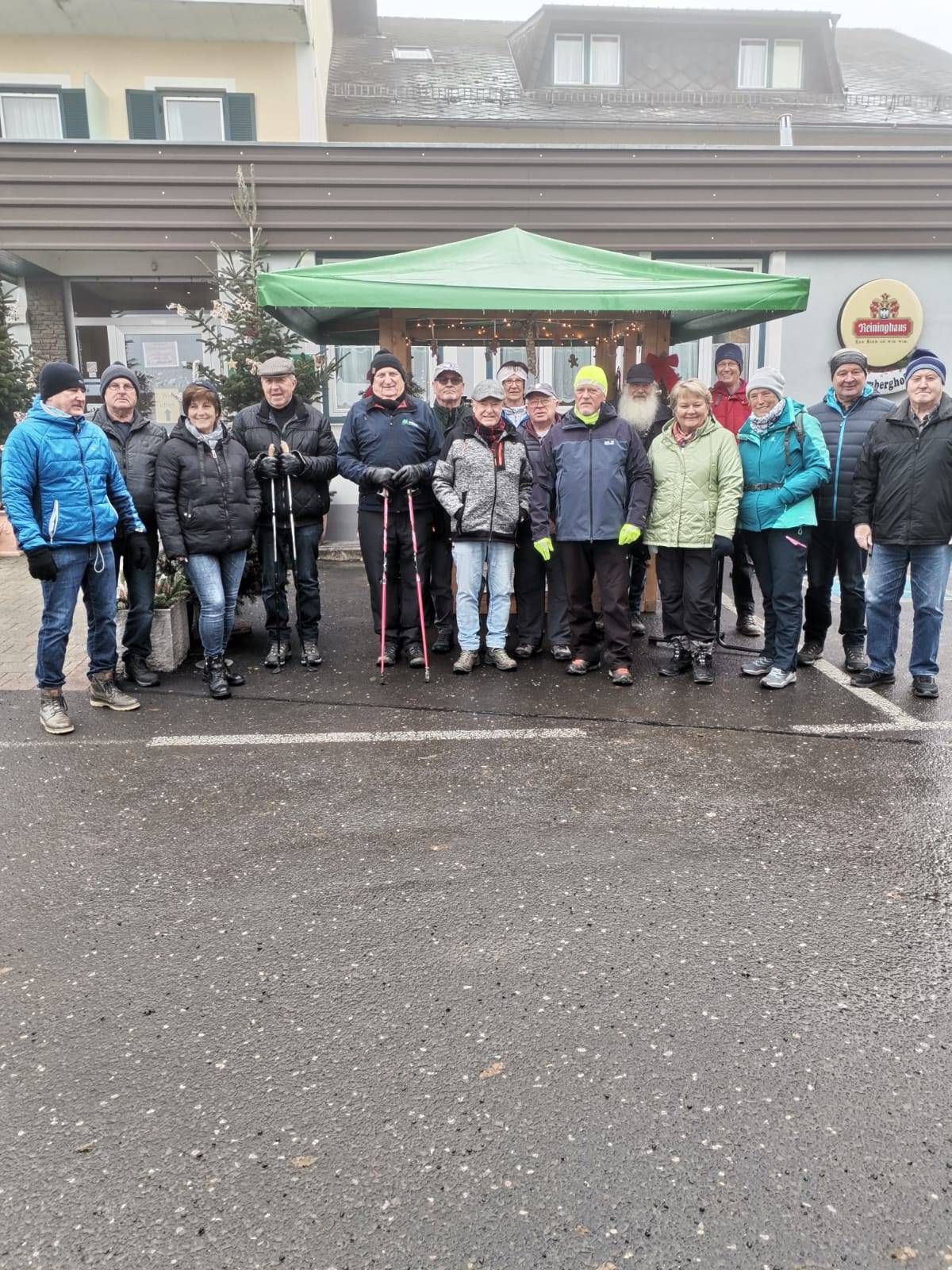 Eine Gruppe von Menschen in Winterkleidung steht auf einem Parkplatz mit einem grünen Vordach hinter ihnen. Es gibt Bäume und ein Gebäude im Hintergrund.