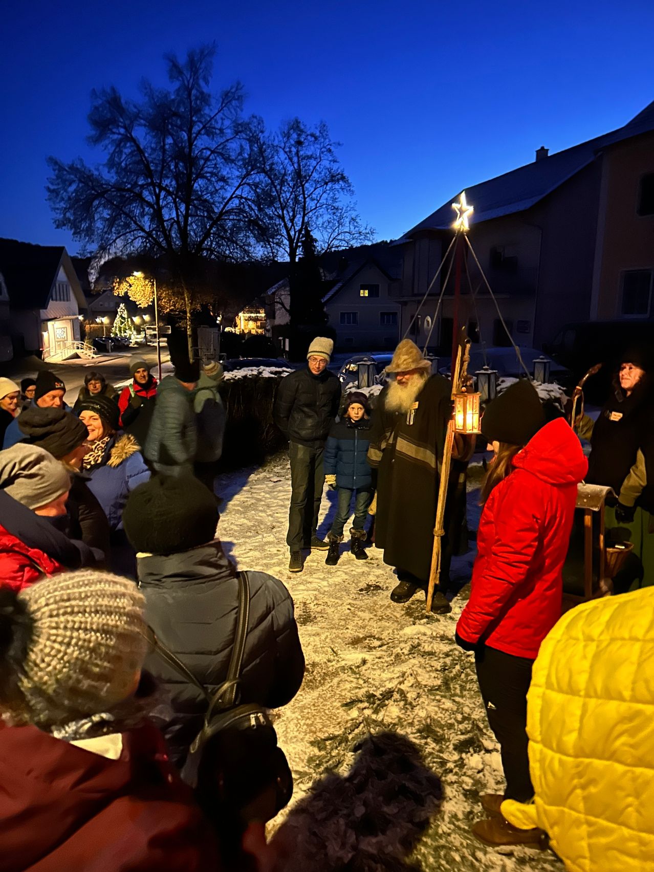 In einem verschneiten Dorf versammeln sich bei Nacht einige Personen um eine Laterne, wobei eine Person als historische Figur gekleidet ist.