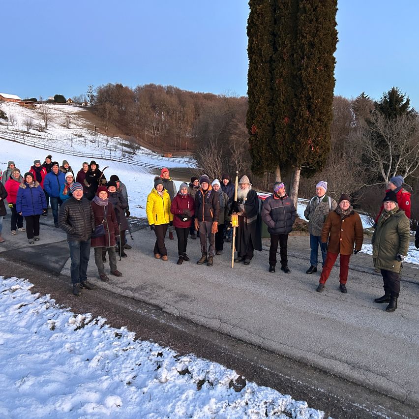 Eine Gruppe von Menschen steht auf einer verschneiten Straße mit einem Baum dahinter, sie tragen Winterkleidung. Einige halten Stöcke. Der Boden ist mit Schnee bedeckt.