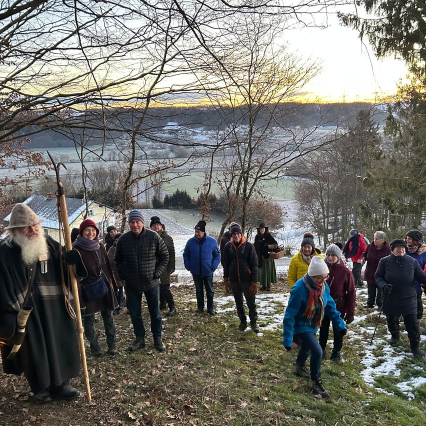 Eine Gruppe von Menschen, gekleidet in Winterkleidung, steht auf einem verschneiten Hügel und blickt in Richtung der untergehenden Sonne, mit Bäumen und einem Haus im Hintergrund.