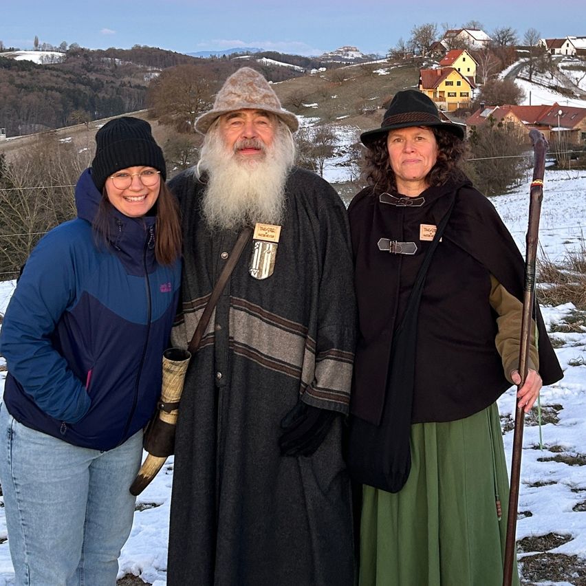 Drei Personen stehen im Schnee. Der Mann hat einen langen Bart und trägt einen Umhang. Die Frauen tragen mittelalterliche Kleidung und halten eine Horn und einen Stab.