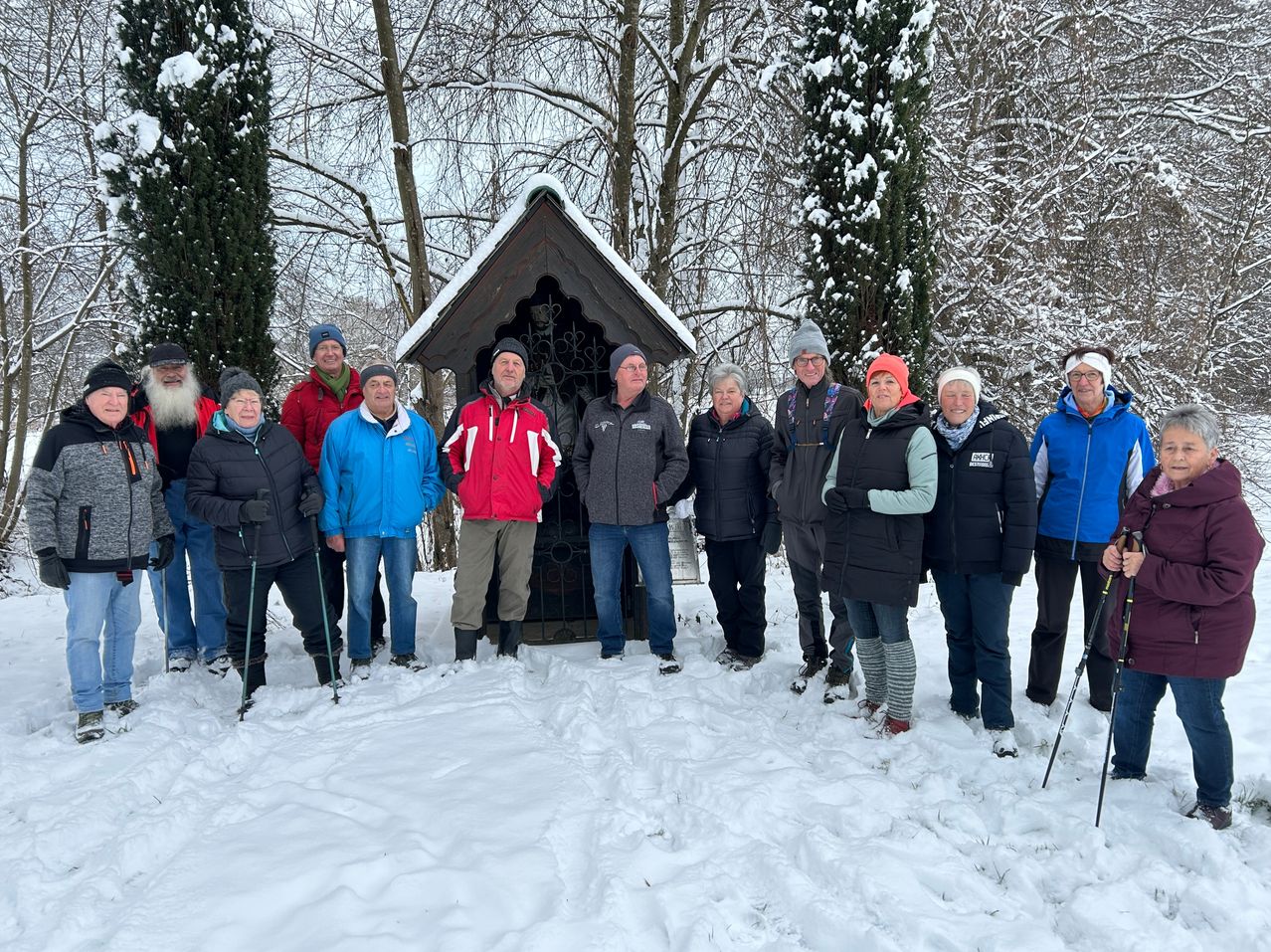Eine Gruppe von Menschen in Winterkleidung steht im Schnee neben einer kleinen Holzkapelle. Sie halten Skistöcke in den Händen und scheinen eine Winterwanderung zu unternehmen.
