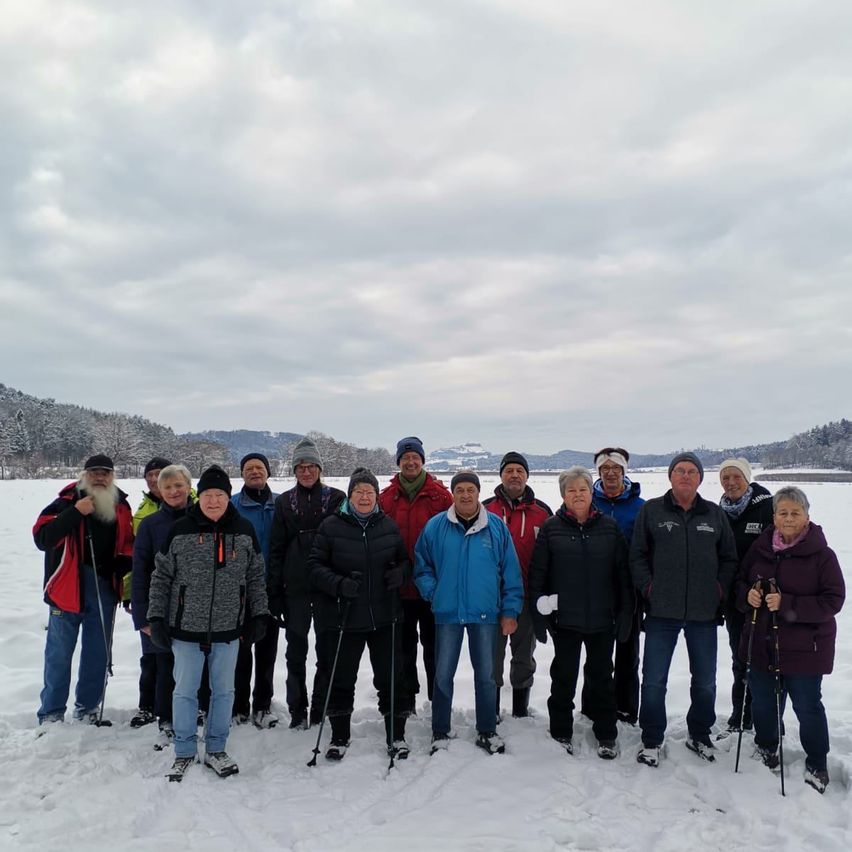 Eine Gruppe von Menschen posiert für ein Foto auf einem verschneiten Feld mit Bergen im Hintergrund. Sie tragen alle Winterkleidung und halten Skistöcke in der Hand.