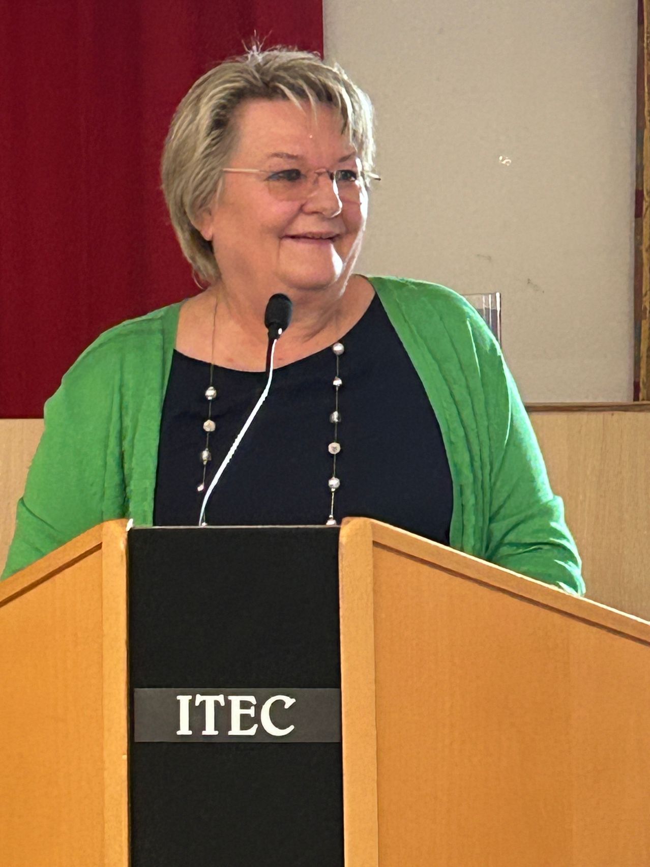 A woman speaks at a podium with a microphone. She wears glasses and a necklace with pearls. Behind her, a red backdrop and a wooden wall are visible. The podium has the letters ITEC.