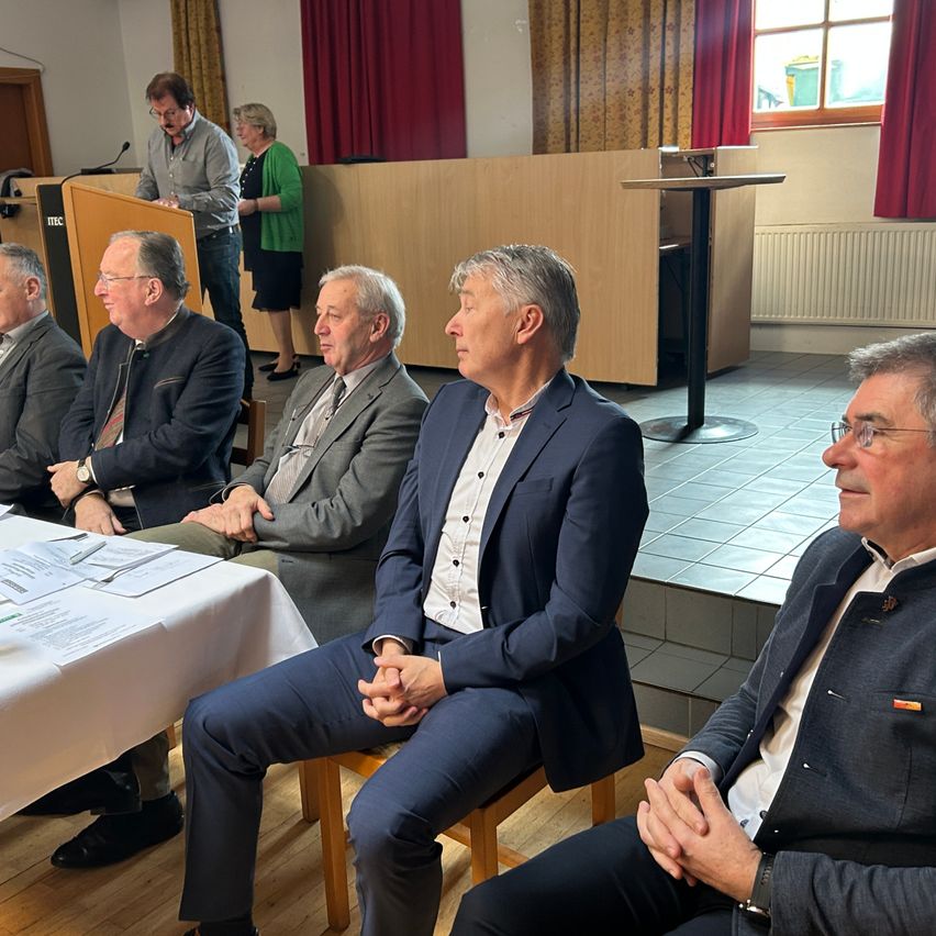 Several men in suits sit around a table, engaged in conversation. A man stands at a podium with a microphone. Behind him, a woman stands, wearing a green sweater. The room has wooden floors and red curtains.
