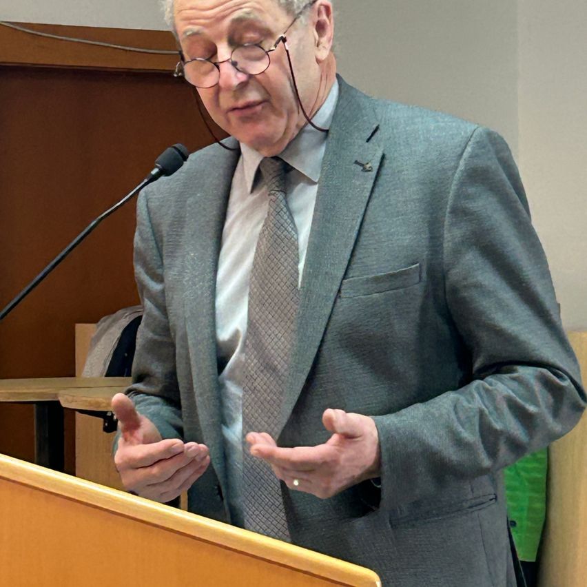 An elderly man wearing a gray suit and tie stands at a podium with a microphone, gesturing while speaking.