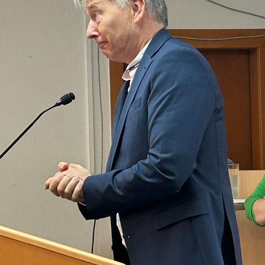 A man is standing at a podium, facing forward, and appears to be giving a speech. He is wearing a blue suit and has a microphone in front of him.