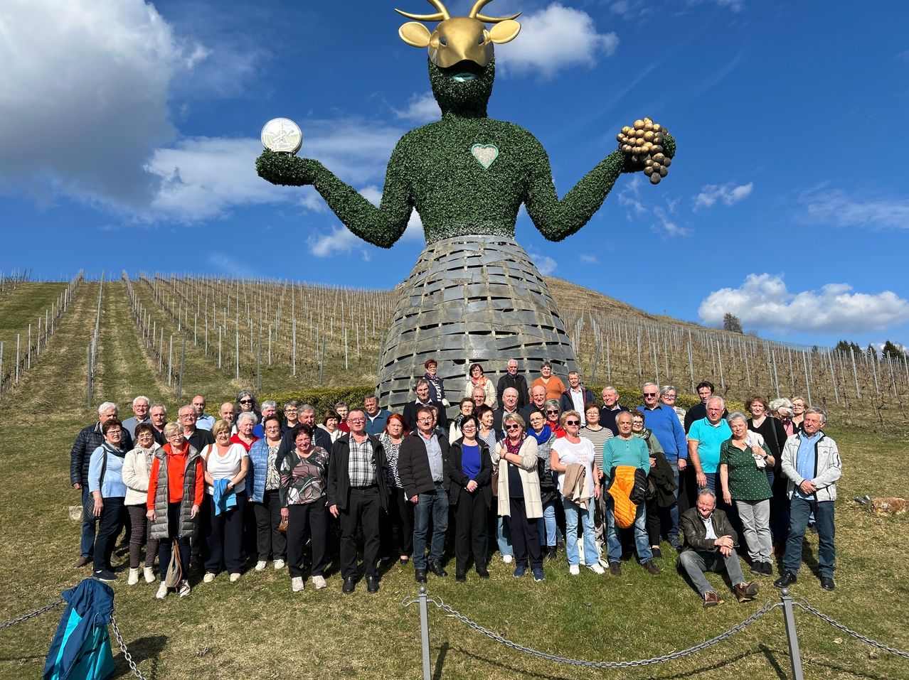 Eine Gruppe von Menschen posiert vor einer großen, menschenförmigen Skulptur aus Grünpflanzen, die einen goldenen Hirschkopf trägt und eine Münze und Trauben hält. Sie stehen in einem Weinberg mit blauem Himmel im Hintergrund.