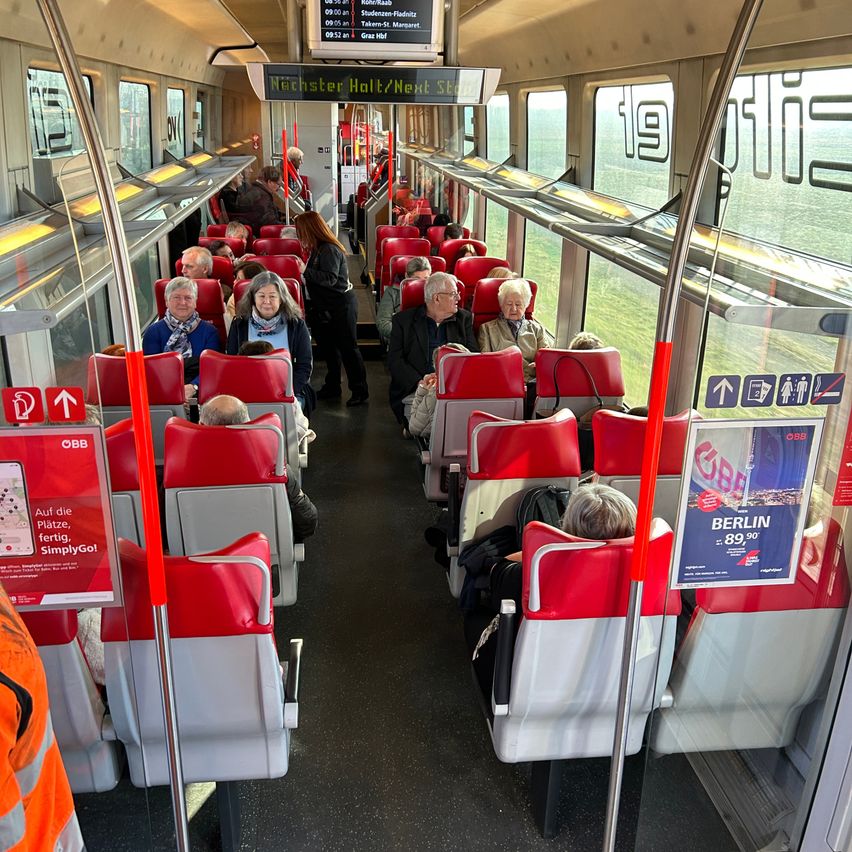 Ein überfüllter Zugwaggon mit roten und weißen Sitzen. Mehrere Passagiere sitzen, während andere stehen oder gehen. Der Zug zeigt Schilder für Ziele und Werbung an. Die Fenster zeigen eine Aussicht auf eine grün bewachsene Landschaft.