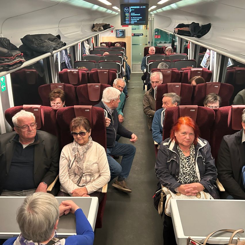 Im Inneren eines Zuges sitzen mehrere ältere Passagiere auf roten Stühlen. Sie schauen umher oder unterhalten sich. Einige tragen Brillen, Schals und Jacken. Taschen hängen an den Gepäckträgern.