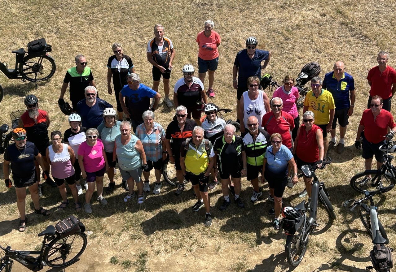 A group of cyclists, all wearing helmets and cycling gear, pose for a photo on a grassy field. Some bikes are visible in the background.