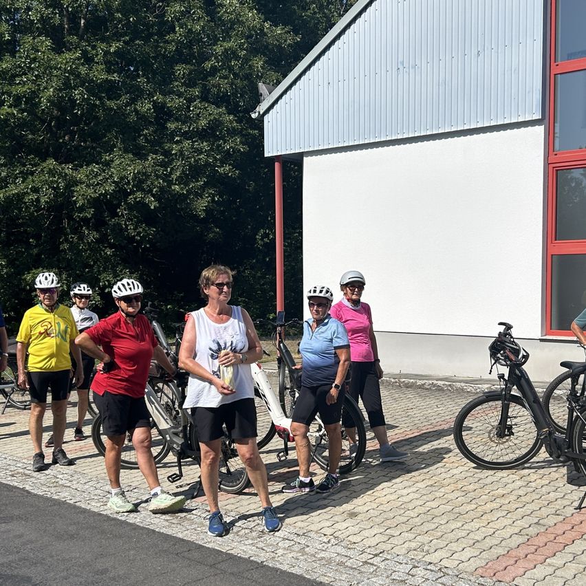 A group of cyclists, all wearing helmets and cycling gear, stand in front of a building with bicycles parked nearby. Trees and a red-framed window are visible in the background.