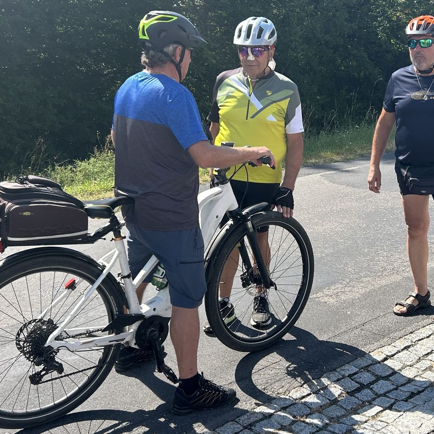 Three men in cycling gear stand on a road, one holding a bicycle. They are wearing helmets and sunglasses. The man on the left holds the handlebars of a white bicycle.
