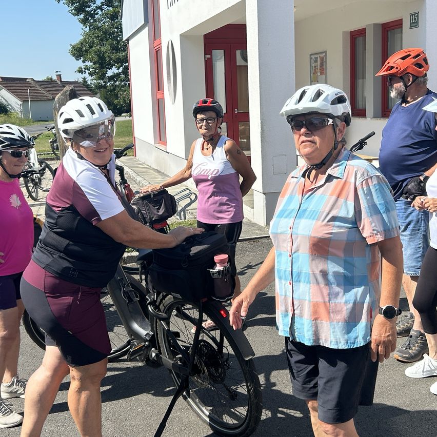 A group of cyclists, wearing helmets and cycling gear, are gathered in front of a building. One woman is holding a bicycle while another woman is holding a bag. A man is standing nearby, wearing a watch.