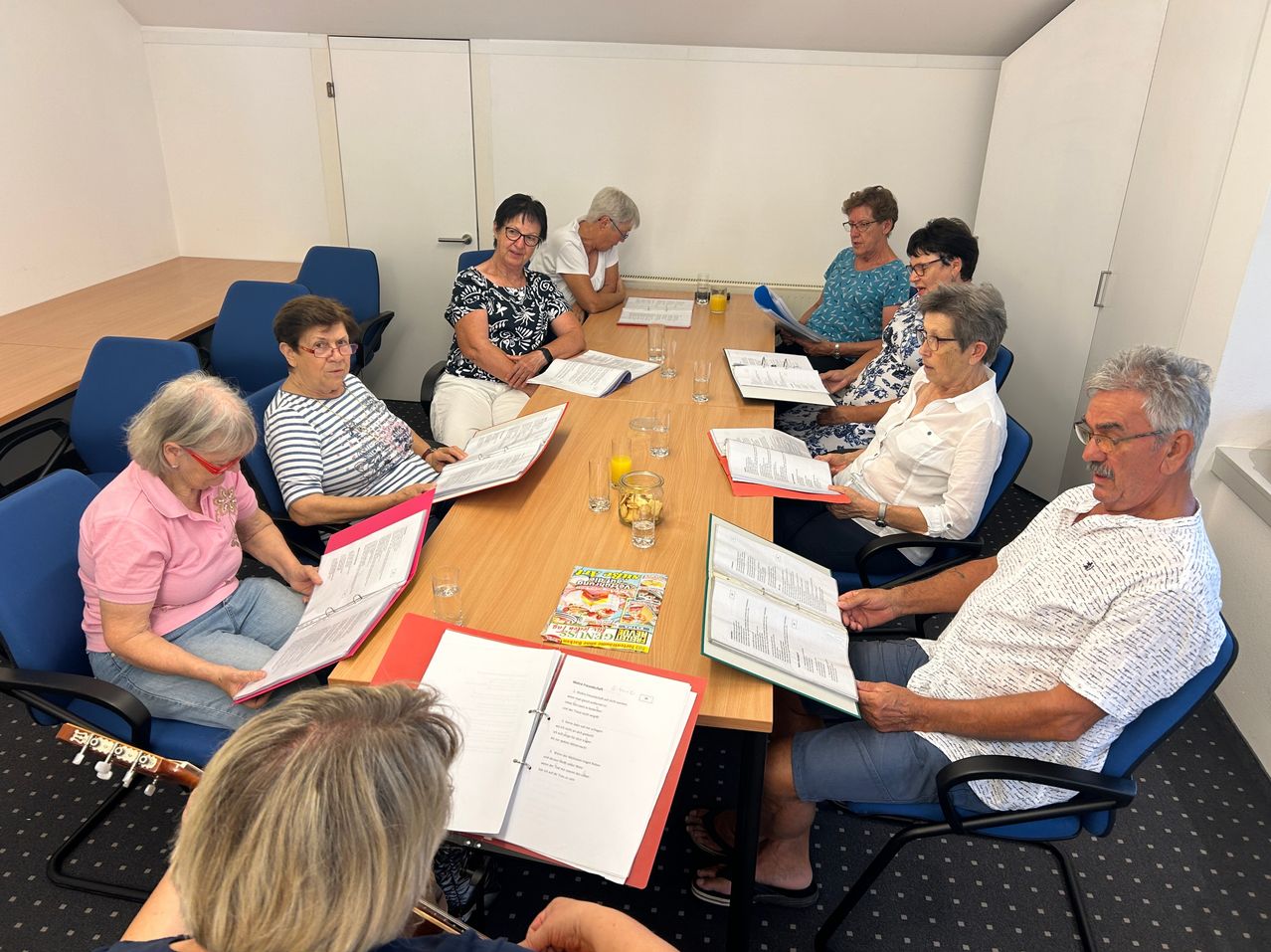 Eine Gruppe älterer Frauen und Männer sitzt um einen Tisch in einem Raum, liest Bücher und Papiere. Auf dem Tisch stehen Gläser, eine Schüssel mit Obst und eine Zeitschrift. Der Raum hat weiße Wände und eine Tür.