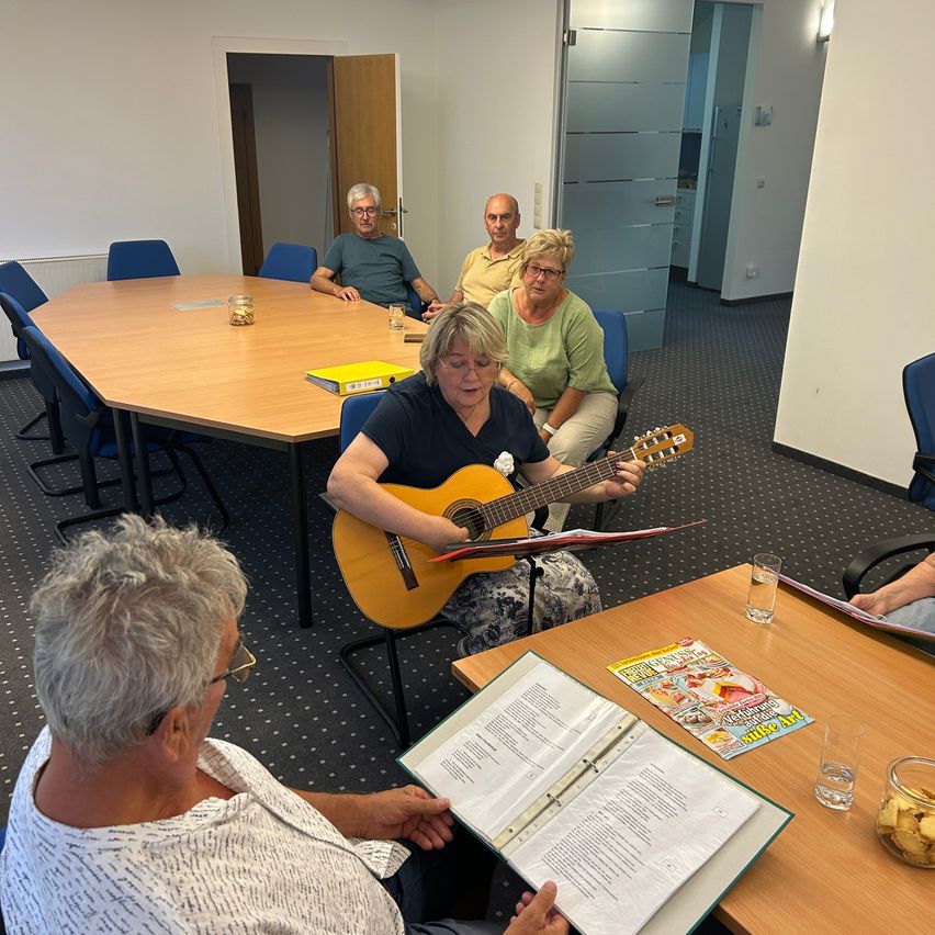 Eine Gruppe älterer Erwachsener sitzt um einen Tisch herum, eine Frau spielt Gitarre, während eine andere Noten liest. Der Raum ist gut beleuchtet mit einem Teppichboden.