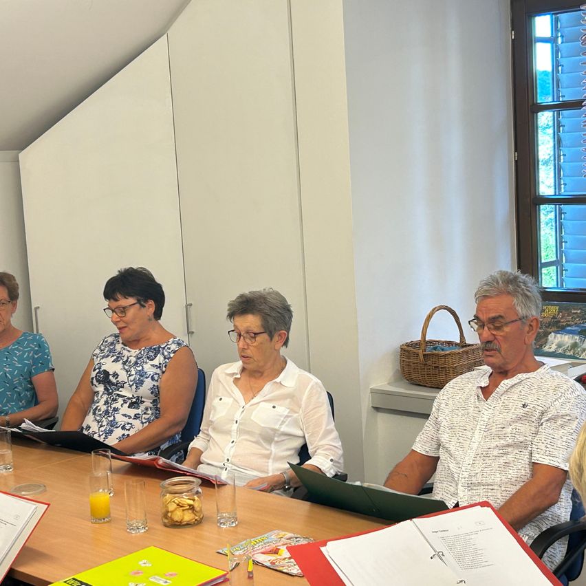 Fünf Personen sitzen um einen Tisch herum in einer Besprechung, mit Papieren, Gläsern und einem Korb auf dem Tisch. Eine Person liest ein Buch.