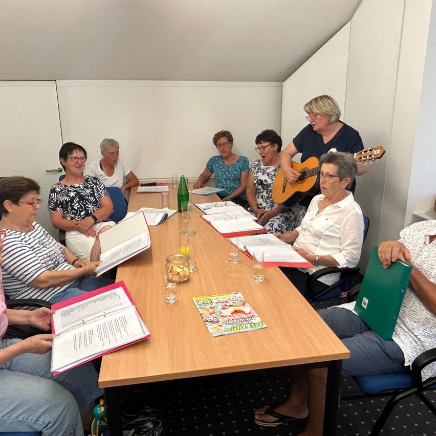 Eine Gruppe von Frauen sitzt um einen Tisch, eine Frau steht und spielt Gitarre, andere lesen aus Büchern und Papieren.