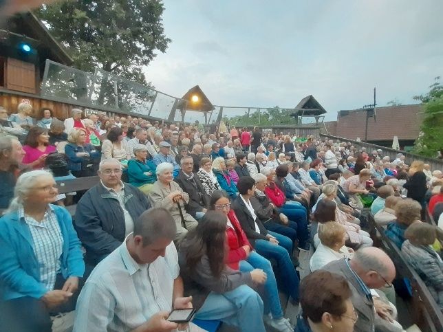 Eine große Menschenmenge sitzt in einem Amphitheater im Freien, einige tragen Brillen und Hüte. Bäume und ein Gebäude sind im Hintergrund sichtbar.