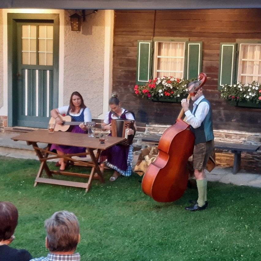 Zwei Frauen sitzen auf einer Bank, eine spielt Gitarre und die andere Akkordeon, während ein Mann Cello vor einem Haus mit Blumen spielt.