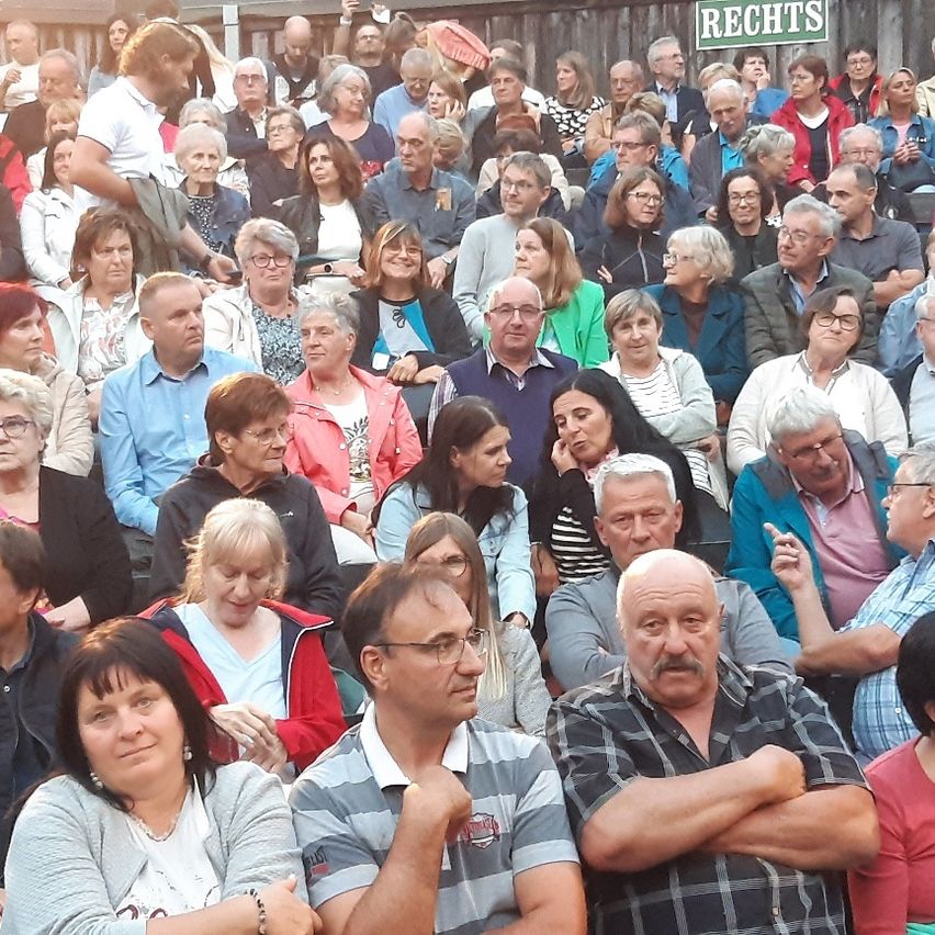 Eine Menschenmenge sitzt in einem Stadion, einige tragen Brillen und Hüte. Ein Schild mit der Aufschrift Rechts ist oben sichtbar.