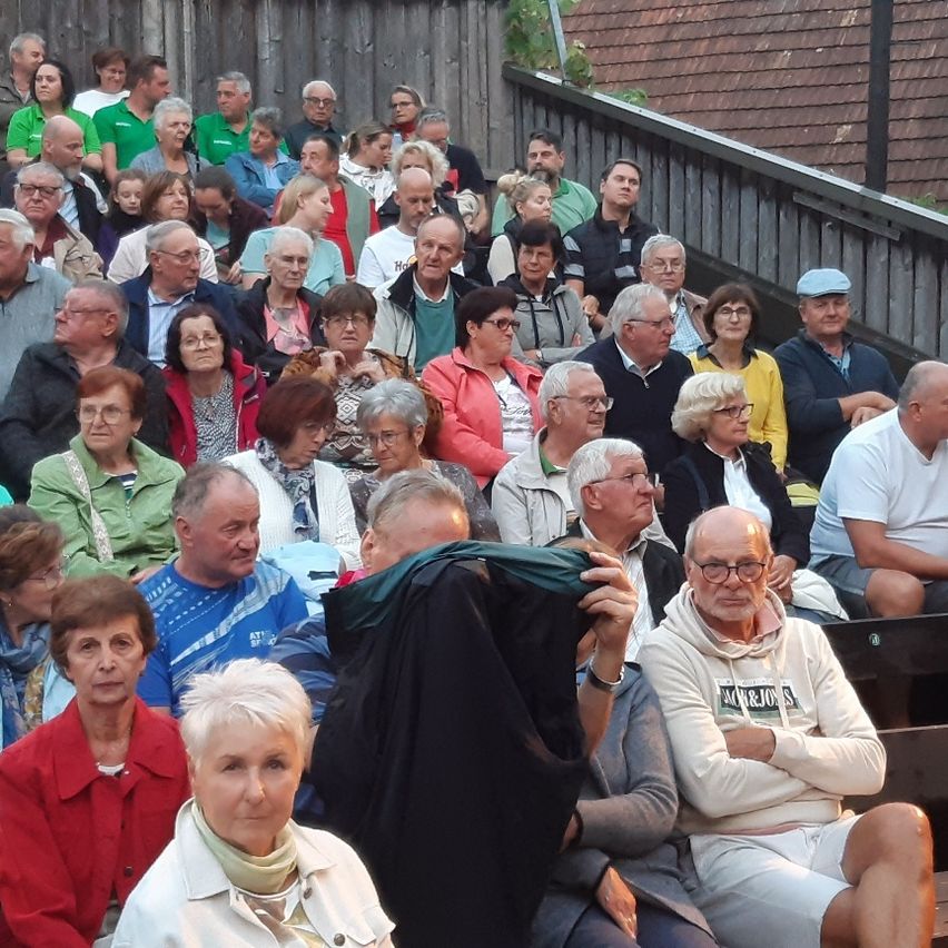 Eine Gruppe älterer Menschen, die meisten mit Brille, sitzen in Reihen auf hölzernen Bänken. Die Person im Vordergrund trägt eine grüne Jacke und ist teilweise durch einen schwarzen Stoff verdeckt.