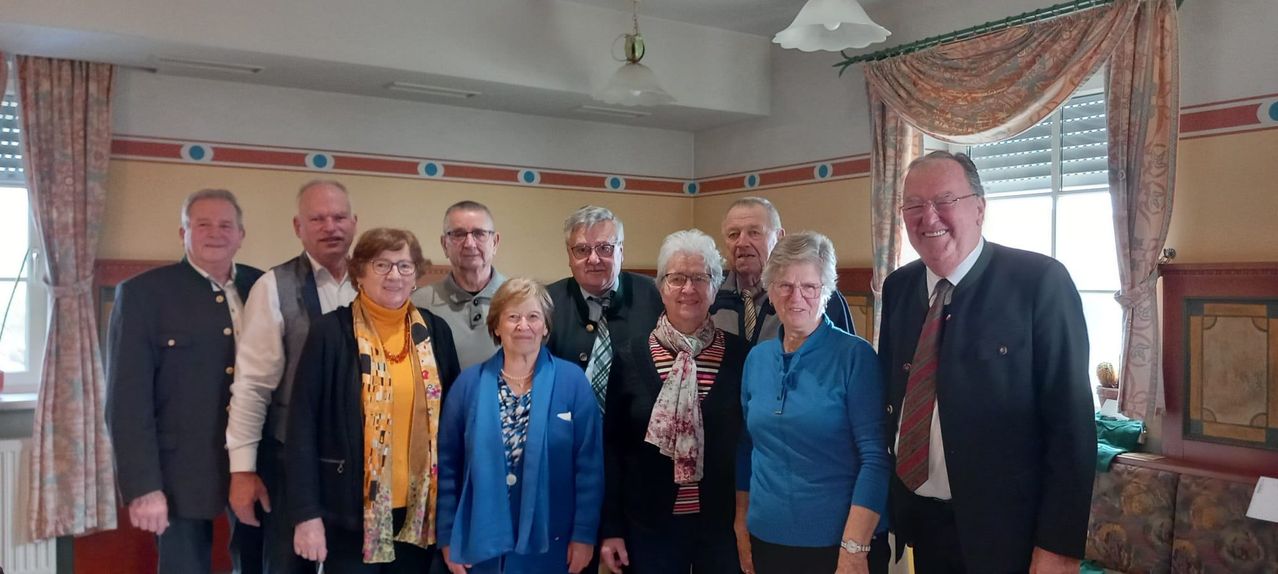 Eine Gruppe älterer Erwachsener steht lächelnd in einem Raum. Sie tragen Winterkleidung und einige Brillen. Der Raum hat dekorative Wandmuster und hängende Lampen.