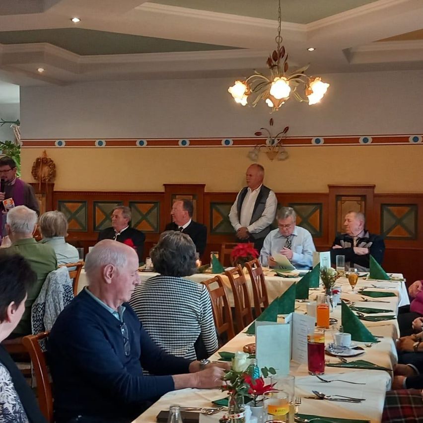 Eine Gruppe älterer Menschen sitzt um einen langen Tisch in einem Speisesaal. Ein Mann steht am Kopfende des Tisches und spricht. Eine Hängelampe hängt von der Decke und ein Kranz ist an der Wand.