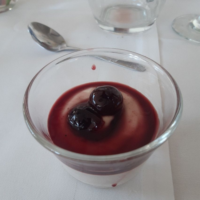 Ein Glas mit roter Kirschsauce und zwei Kirschen darauf steht auf einer weißen Tischdecke. Ein Löffel und ein Glas befinden sich hinter dem Glas.