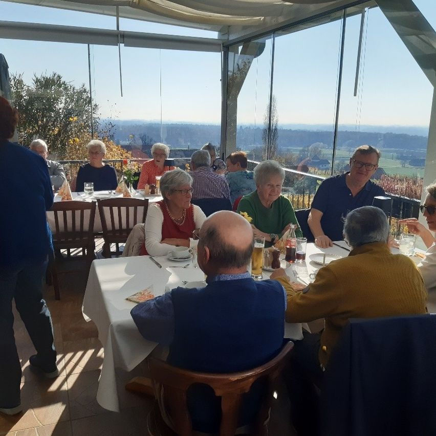 Eine Gruppe älterer Menschen sitzt in einem Restaurant mit Blick auf die Landschaft um einen Tisch herum. Einige von ihnen tragen Brillen und Halsketten. Auf dem Tisch stehen Gläser, Teller und eine Blumenvase.