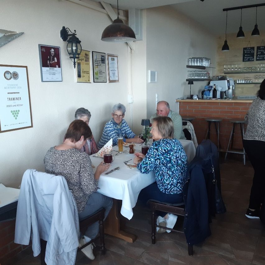 Eine Gruppe von Menschen sitzt an einem Tisch in einem Restaurant, trinkt und unterhält sich. Es gibt Schilder und Lampen an der Wand und eine Theke mit Flaschen.