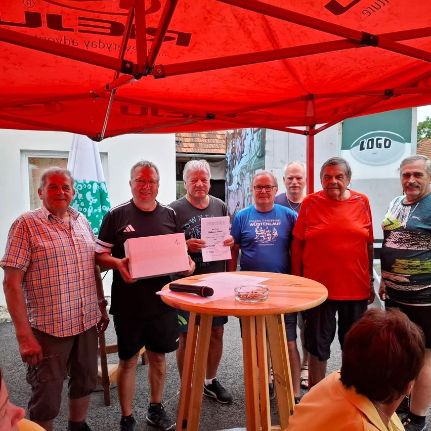 Sieben Männer stehen unter einem roten Baldachin, einer hält ein Zertifikat, andere lächeln. Ein kleiner Tisch mit Mikrofon und Aschenbecher steht davor. Eine Flagge und ein Gebäude sind im Hintergrund.