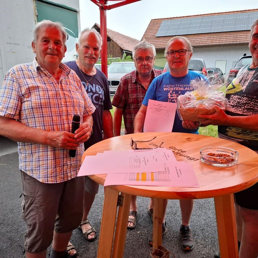 Fünf Männer stehen neben einem Holztisch und halten ein Mikrofon, Papiere und einen Geschenkkorb. Dahinter sind Solarmodule auf dem Dach eines Hauses montiert.