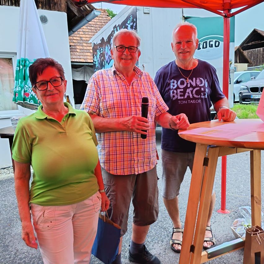 Drei Menschen stehen unter einem orangefarbenen Sonnenschirm. Eine Frau hält eine blaue Tasche und zwei Männer ein Mikrofon. Der Mann rechts trägt ein blaues T-Shirt mit weißem Text.