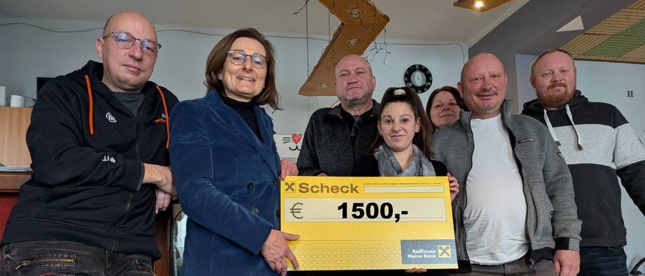 Eine Gruppe von Menschen steht in einem Raum, hält einen großen Scheck über 1500 Euro und posiert für ein Foto.