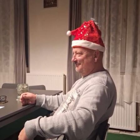 Ein älterer Mann mit einem Weihnachtsmannhut sitzt an einem Tisch mit einem Glas Wasser, lächelt und ist von einem Raum mit Vorhängen und einem Heizkörper umgeben.