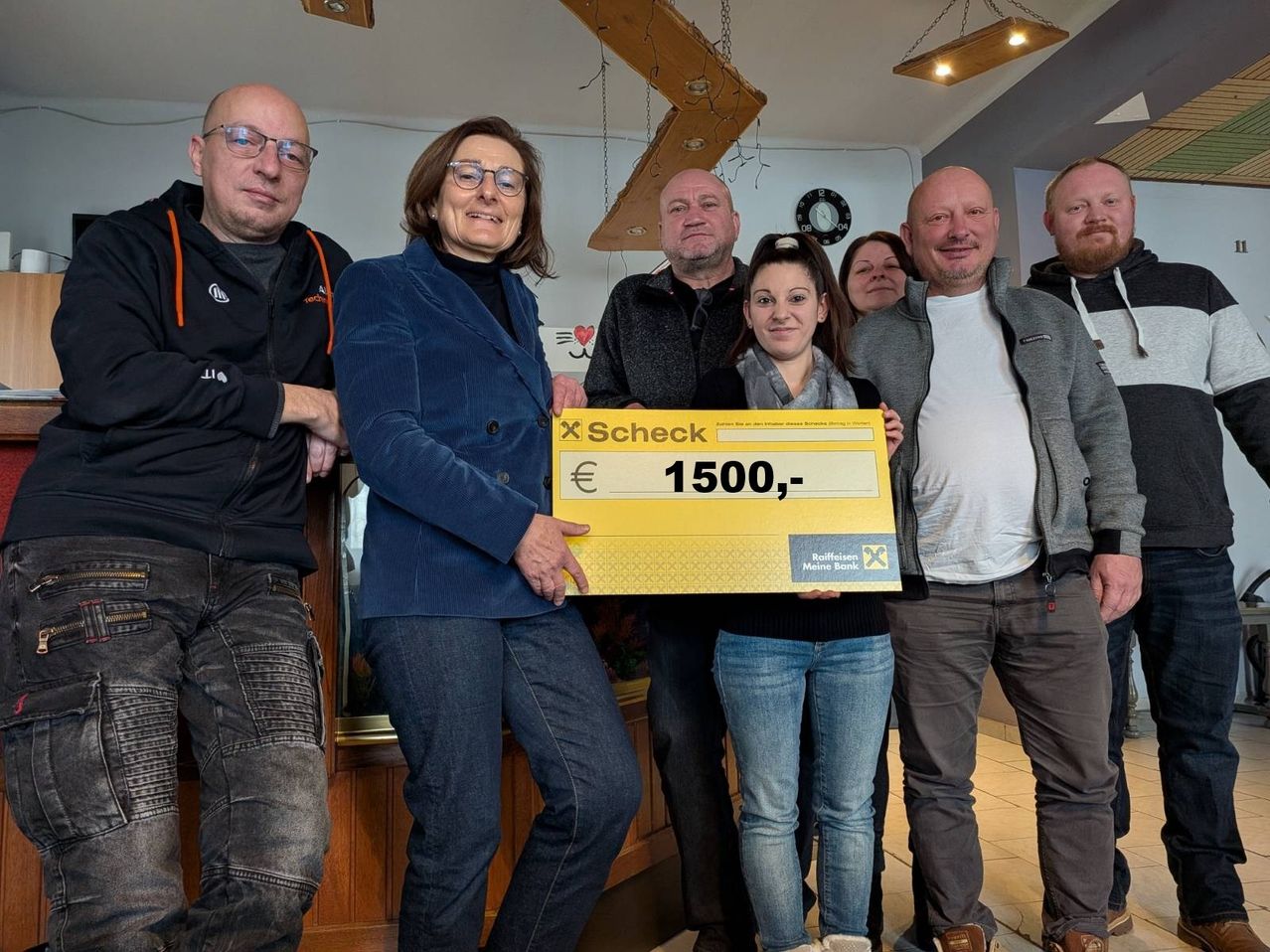 A group of people stands together in a room. A woman holds a large check for 1500 euros. The people behind her look happy and are smiling.