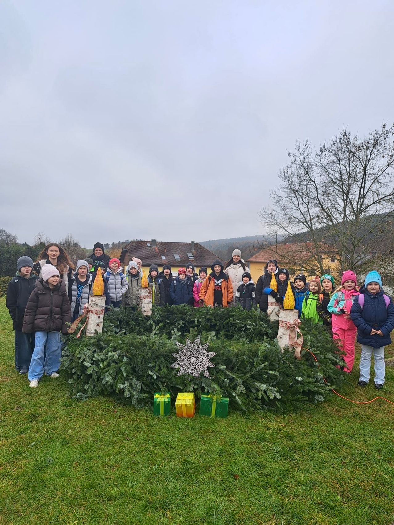 Eine Gruppe von Kindern und Erwachsenen steht um einen Kranz aus immergrünen Pflanzen. Sie tragen Winterkleidung. Hinter ihnen stehen Gebäude und ein Baum. Geschenke liegen vor dem Kranz.