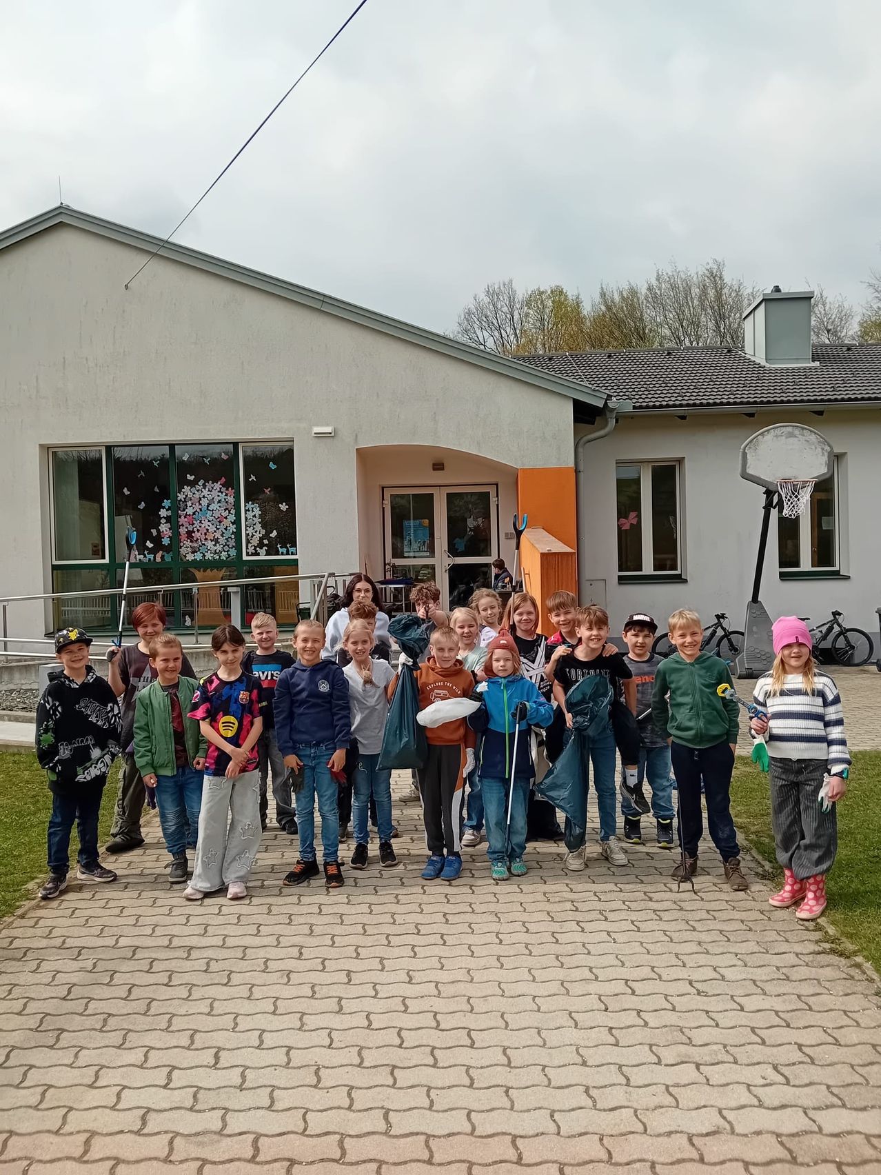 A group of children stands in front of a house with a basketball hoop, some holding cleaning tools. They are on a brick pathway with bicycles parked behind them.