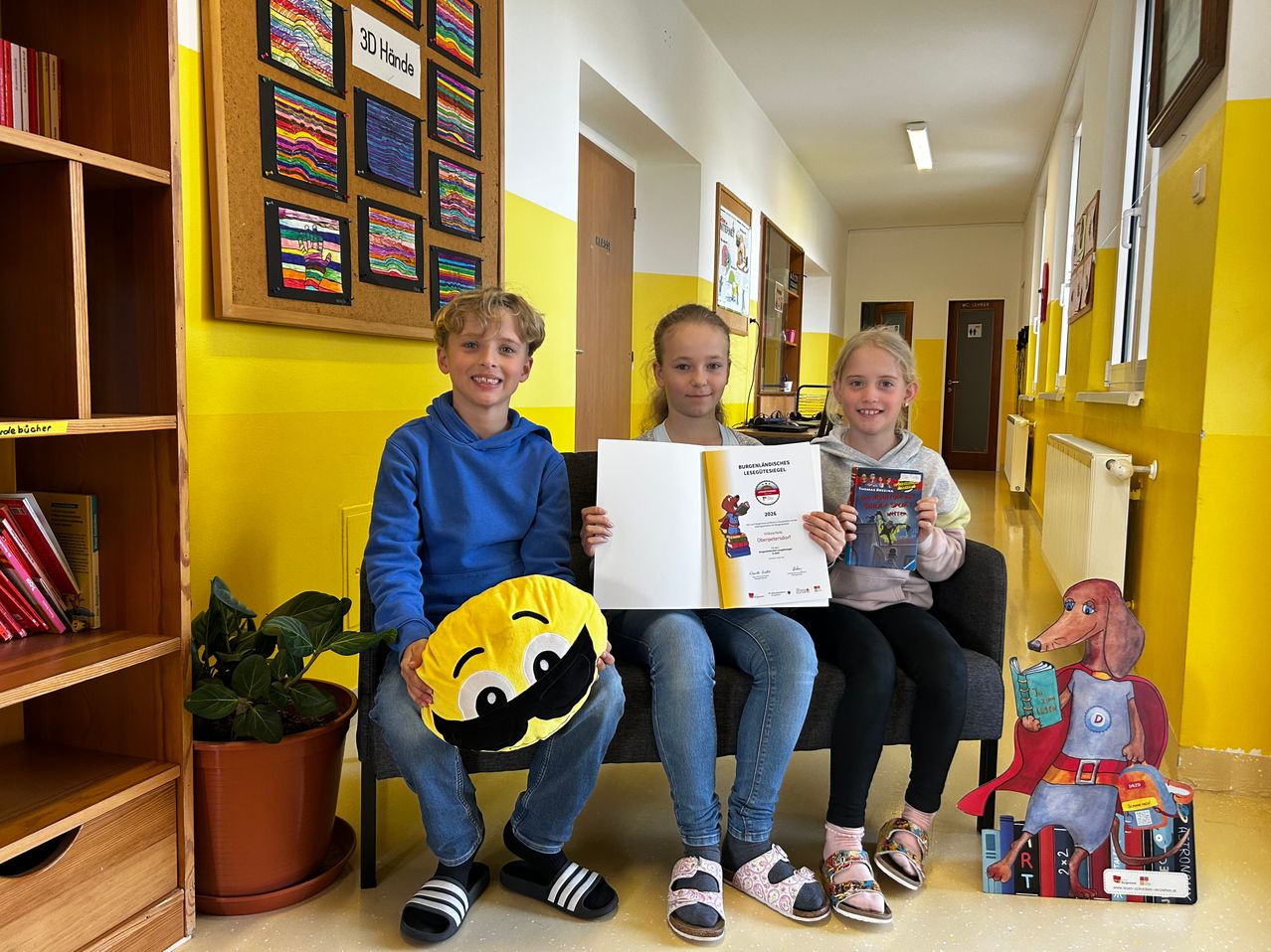 Three children sit on a bench in a yellow hallway, each holding books and smiling. The boy on the left holds a pillow with a smiley face. Behind them is a yellow wall with artwork.