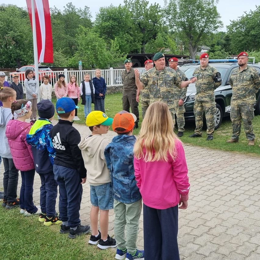 A line of children in casual clothes stand in front of a group of soldiers in camouflage uniforms. A flag is behind them, and a car is parked nearby.