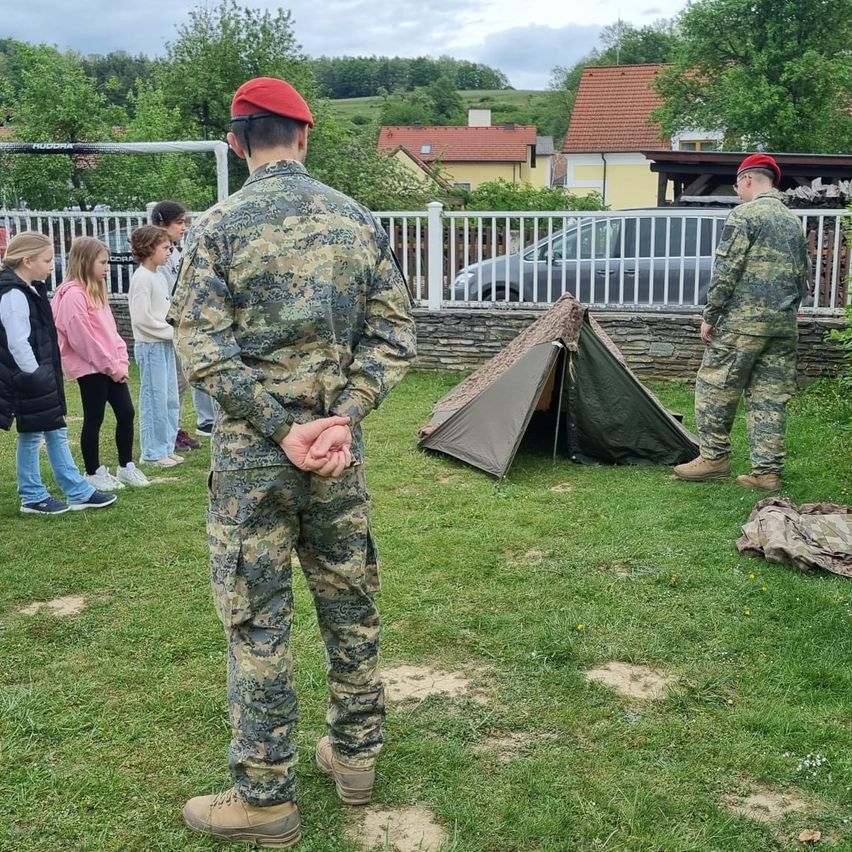 Two soldiers in camouflage stand near a tent on a grass field. Several people, including children, are standing behind a white fence. A car is parked in the distance.